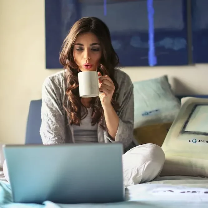 long brown hair woman drinking coffee on laptop sitting on bed